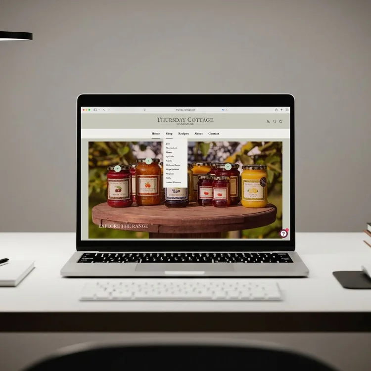Open laptop on a white desk displaying a website for Thursday Cottage with jars of jam and marmalade on a wooden surface